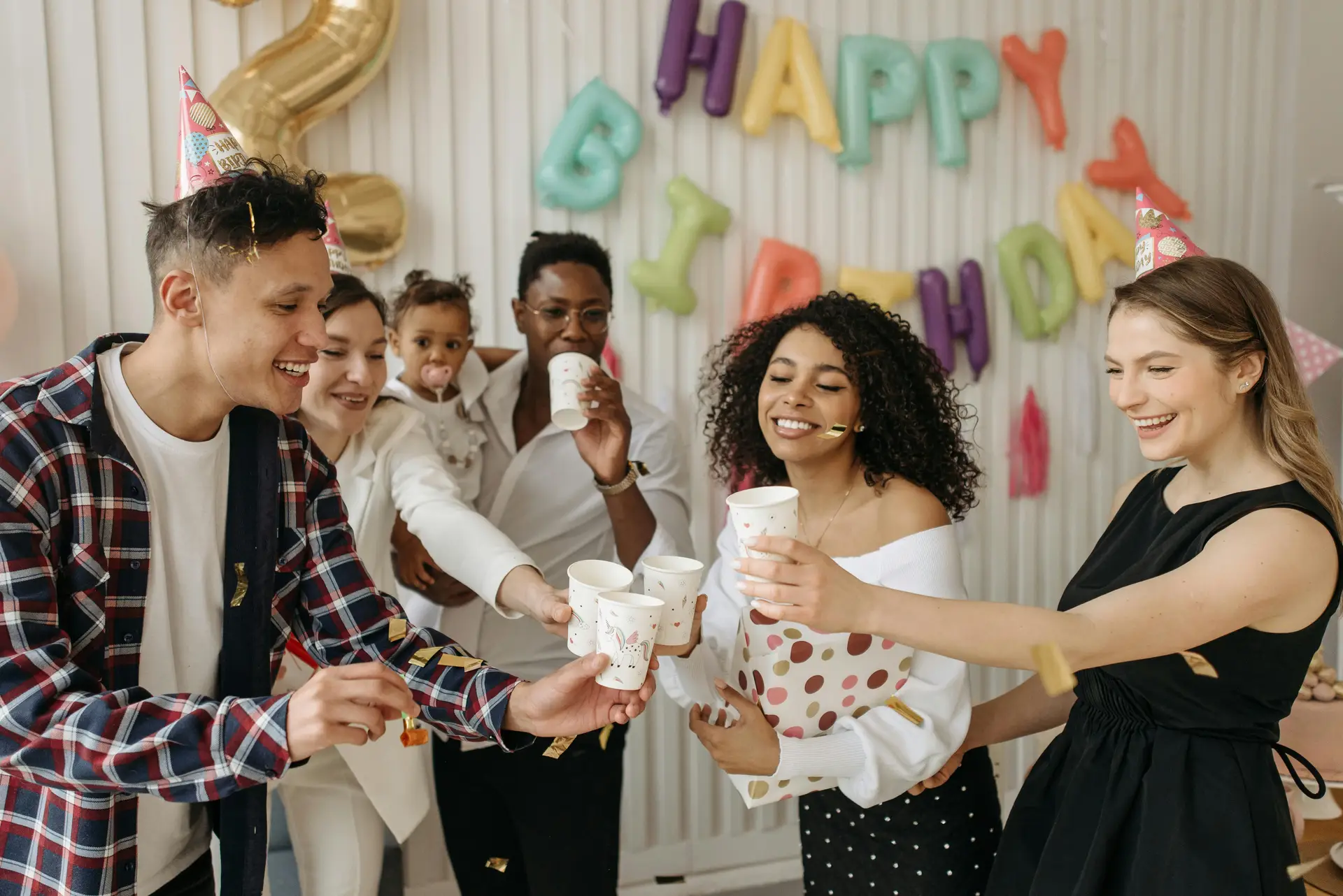 A diverse group of friends celebrating a birthday indoors with party hats and balloons.
